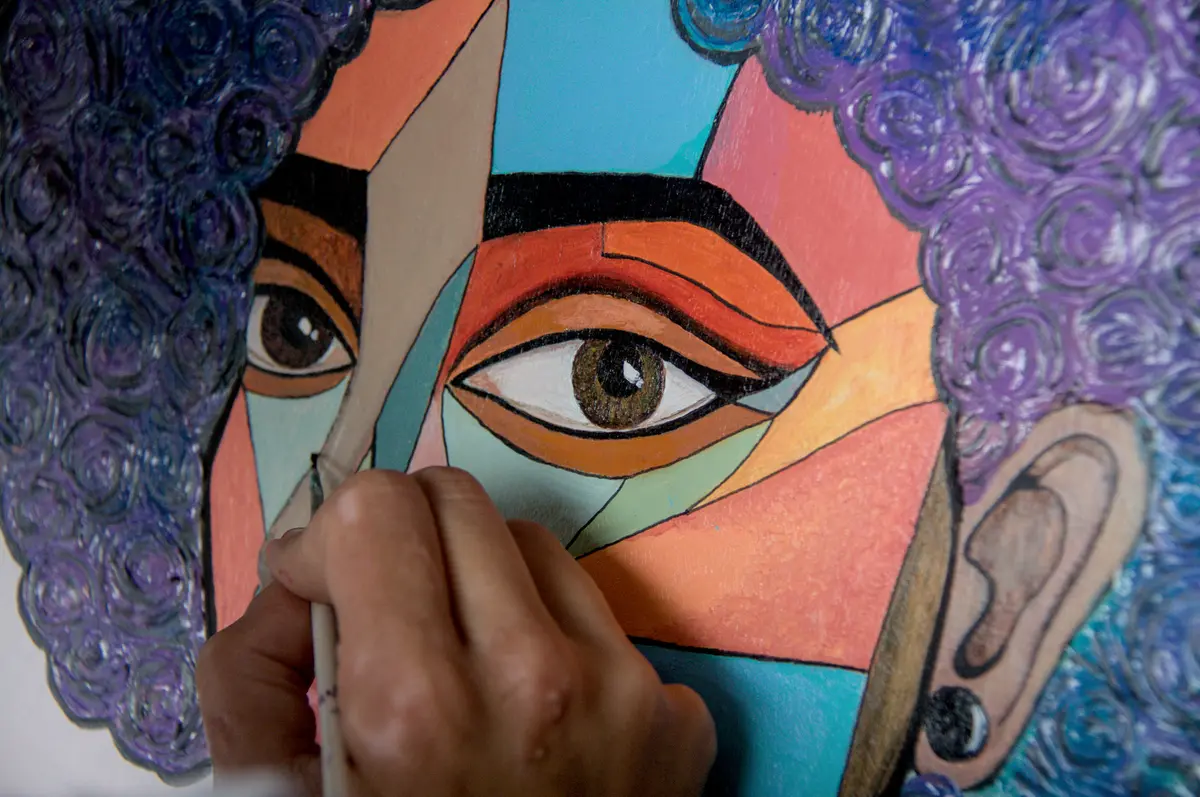 Primer plano de la mano de Najlae pintando un rostro geométrico en corales, turquesas y violetas.